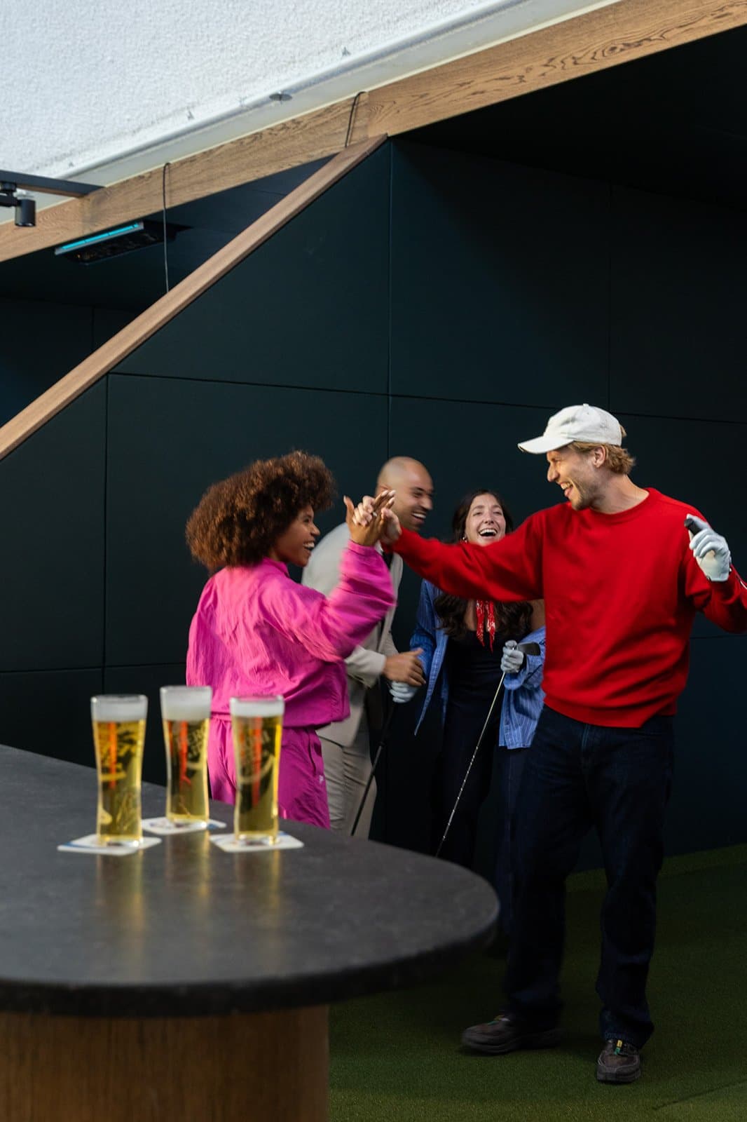 Vieren vier vrienden die lachen en elkaar high-fiven tijdens het spelen van golf in een indoorsportfaciliteit met vier glazen bier op de tafel.