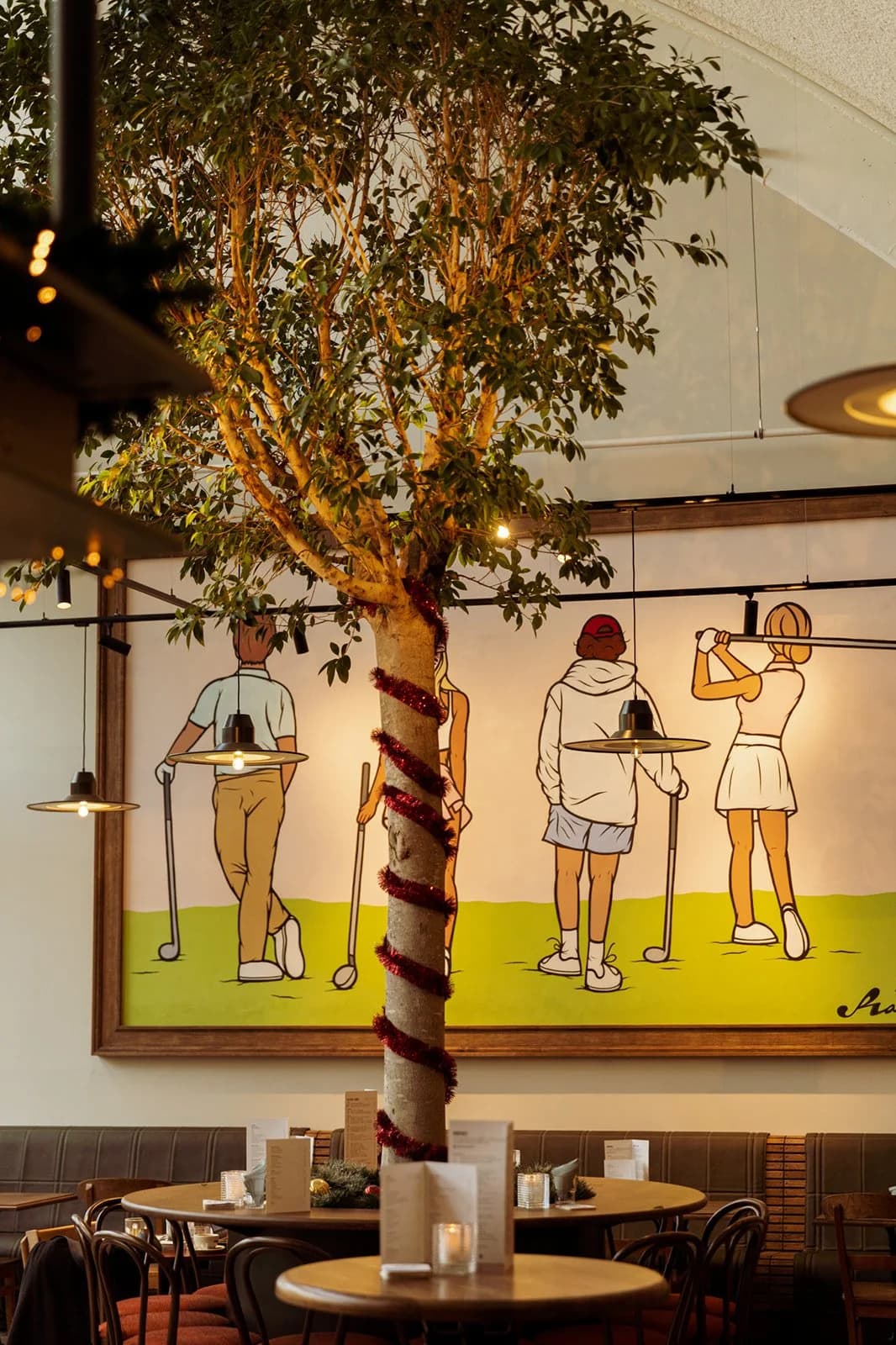 Interieur van een restaurant met een kunstwerk van vier cartoonische figuren die golfen, een kunstboom met lampen en kerstversiering op de stam.